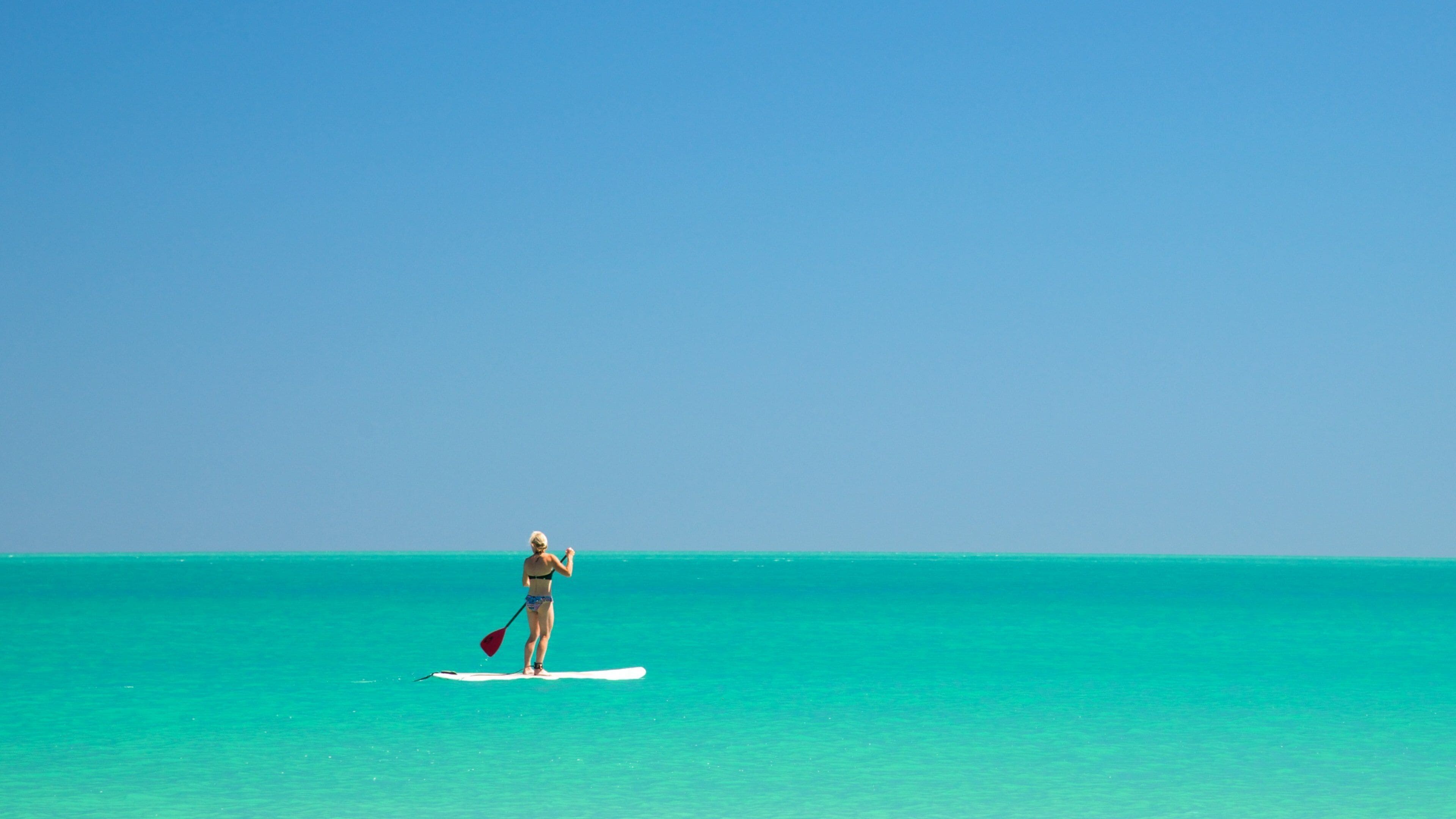 Eine Person steht auf einem Paddleboard auf türkisfarbenem Wasser, der Himmel ist klar und blau.