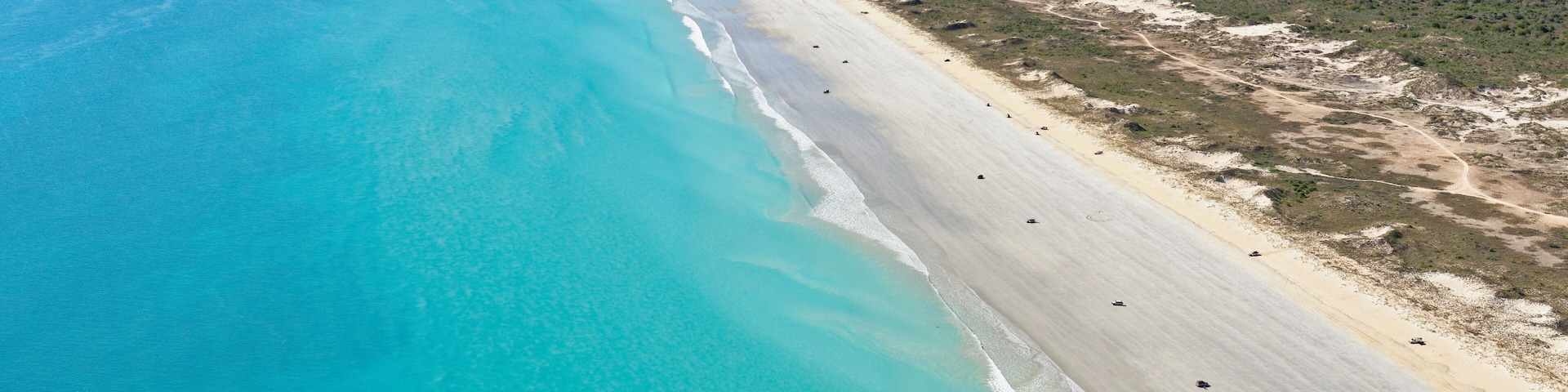 A coastal landscape with a sandy beach, clear turquoise waters, and a distant forested area.