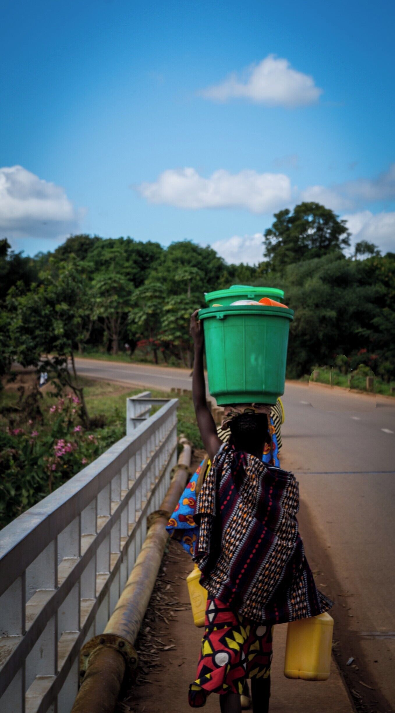 A person carrying a green container on their head, walking along a road with a metal railing on one side and trees in the background.