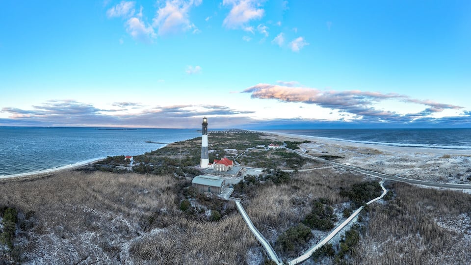 A lighthouse on a coastal bluff with a road leading to it, surrounded by coastal vegetation and a view of the ocean.