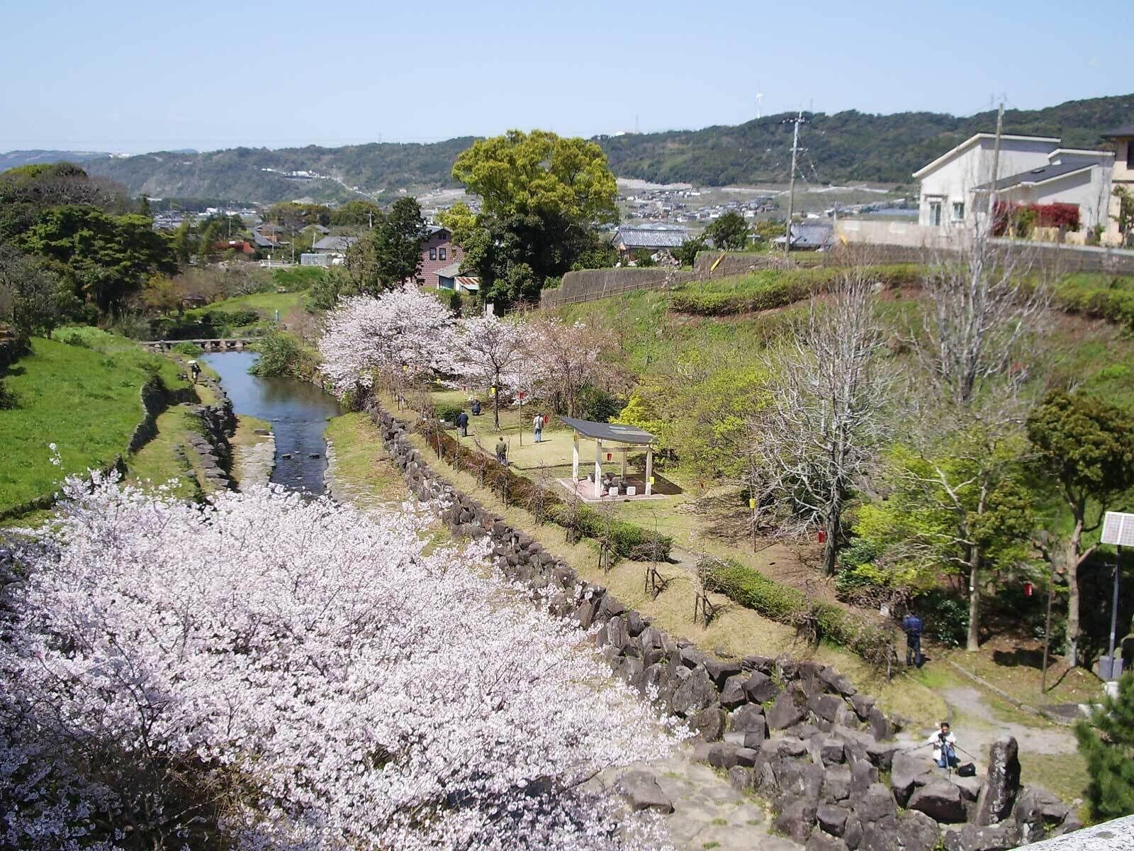 Um rio com cerejeiras em flor, um muro de pedra e um pequeno pavilhão.