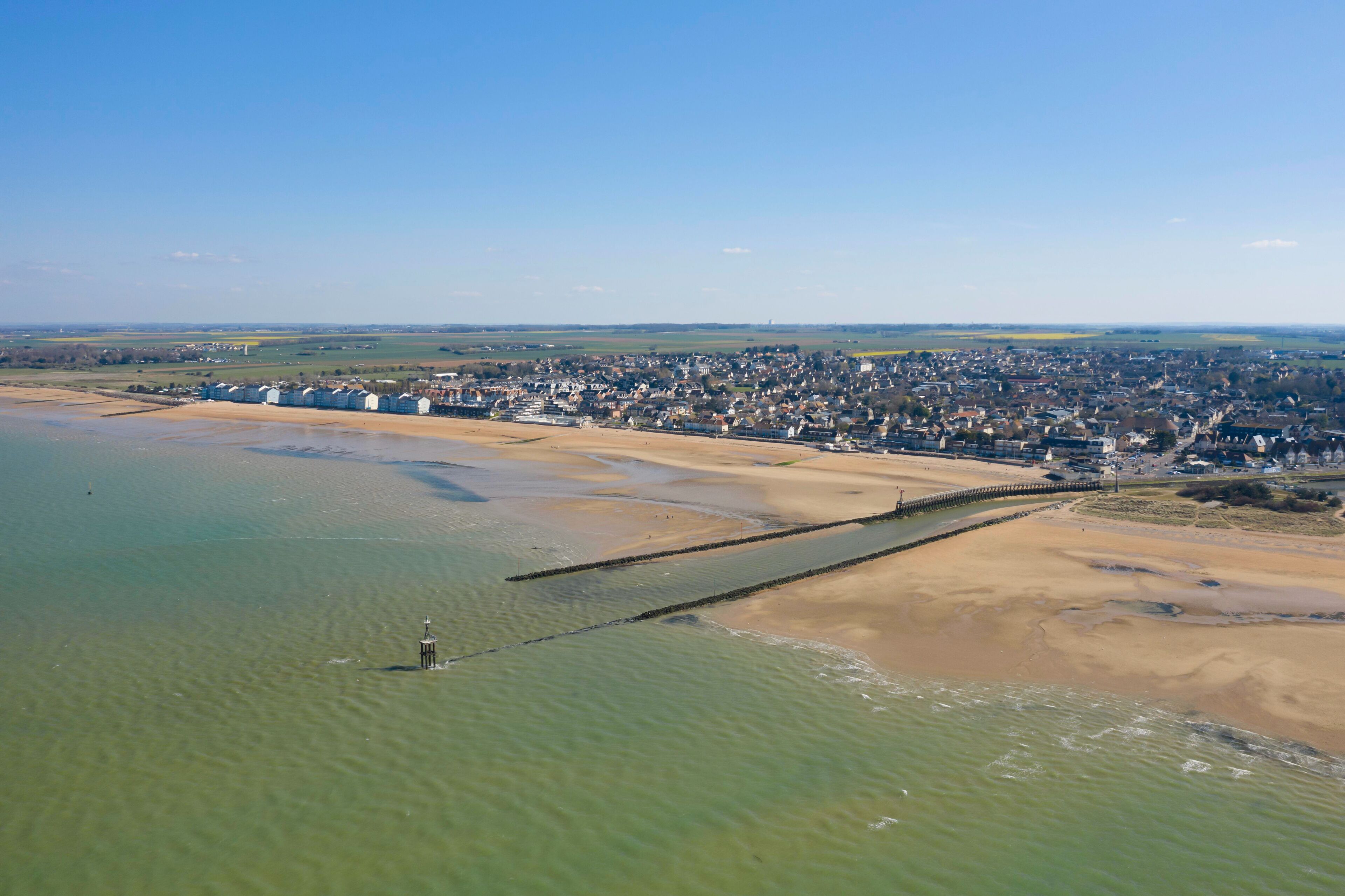 An aerial view of a coastal town with a sandy beach, a pier, and a clear blue sky.