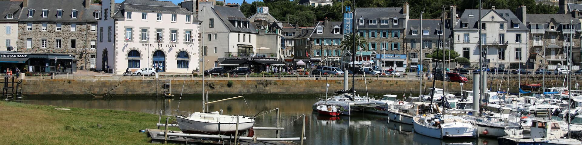 A marina with boats docked, a prominent church with a tower, and a hotel named 'Hotel de Ville' in a coastal town.