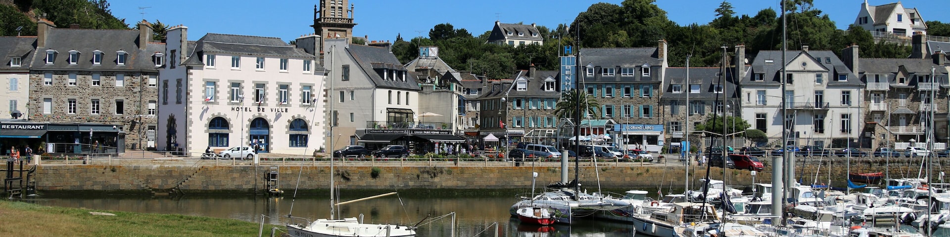 Uma marina com barcos atracados, uma igreja imponente com torre e um hotel chamado 'Hotel de Ville' numa cidade costeira.