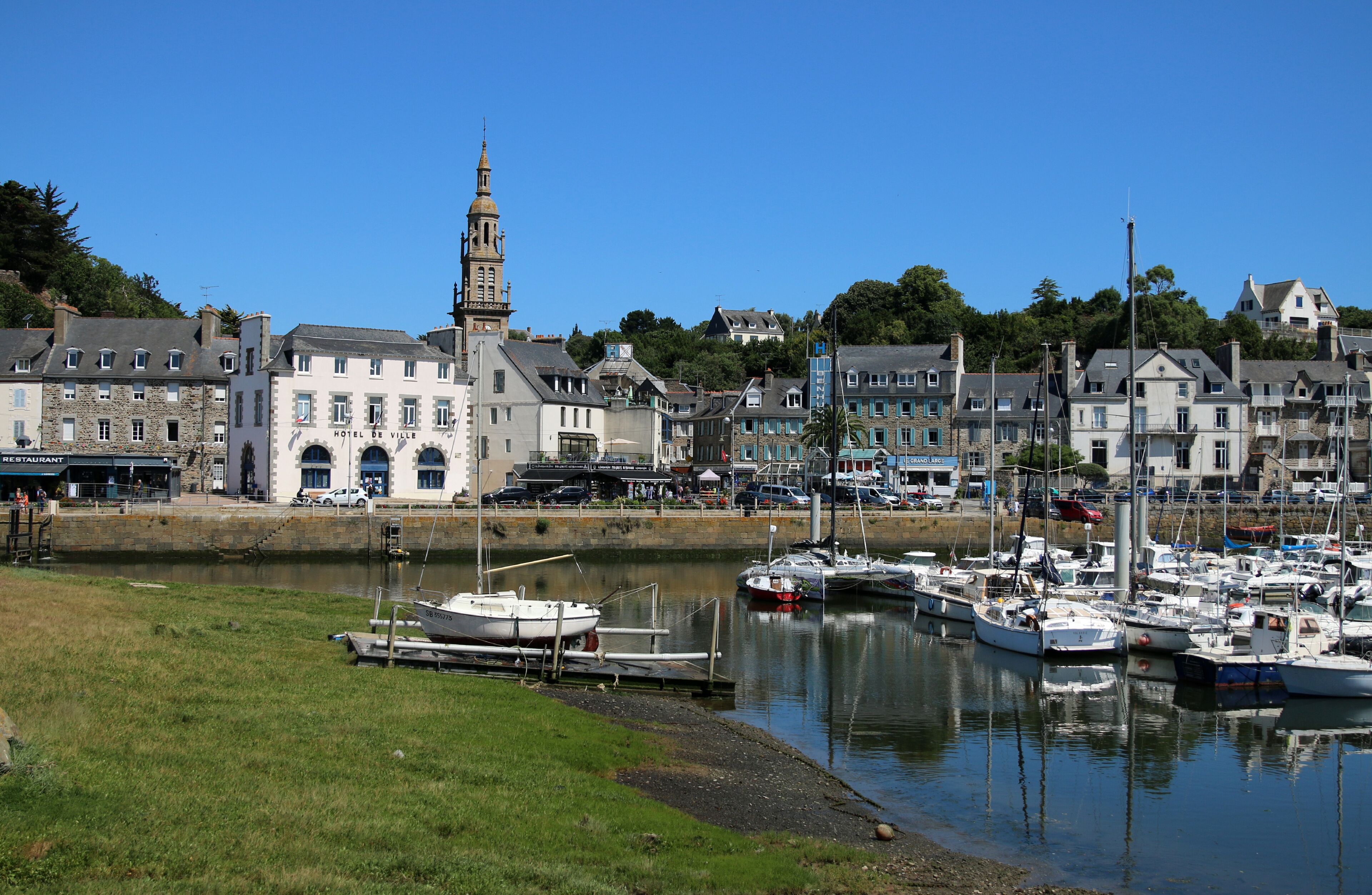 A marina with boats docked, a prominent church with a tower, and a hotel named 'Hotel de Ville' in a coastal town.