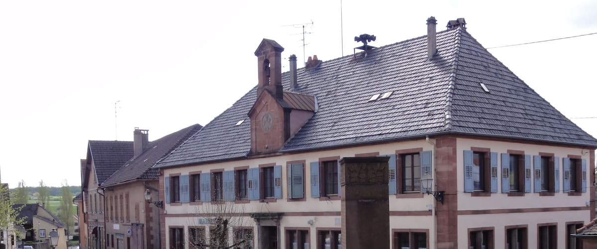 A traditional European building with a steeply pitched roof, multiple windows with shutters, and a small tree in the foreground.