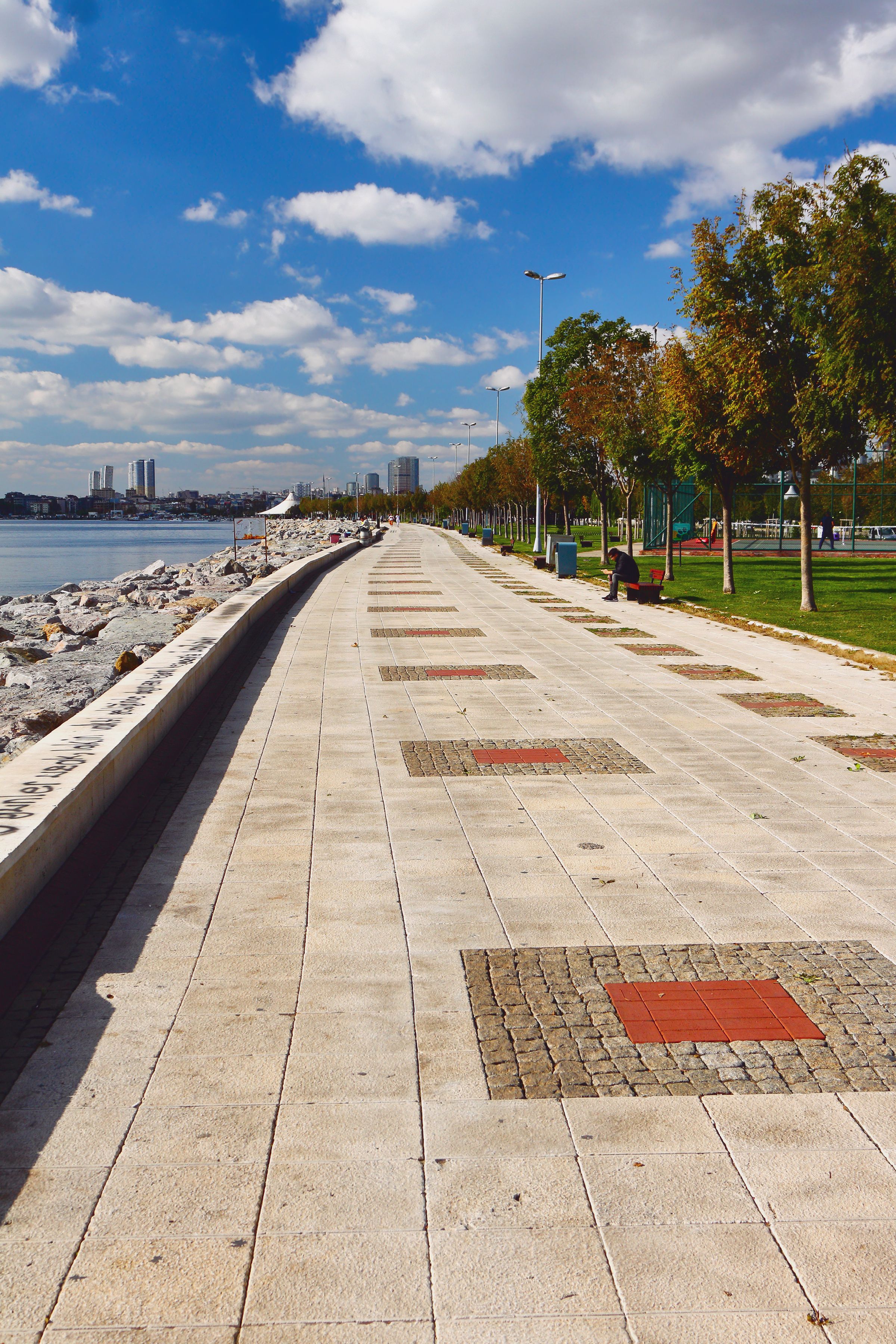 A waterfront promenade with a paved walkway, trees, and a distant city skyline.