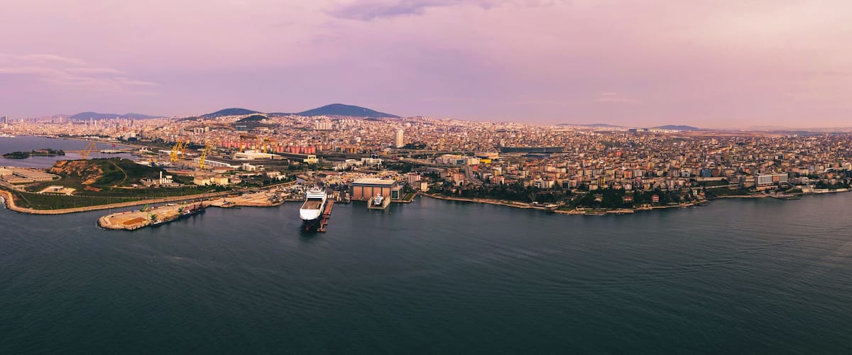 A coastal cityscape with a prominent ship docked at a pier, surrounded by dense urban development and a backdrop of hills.