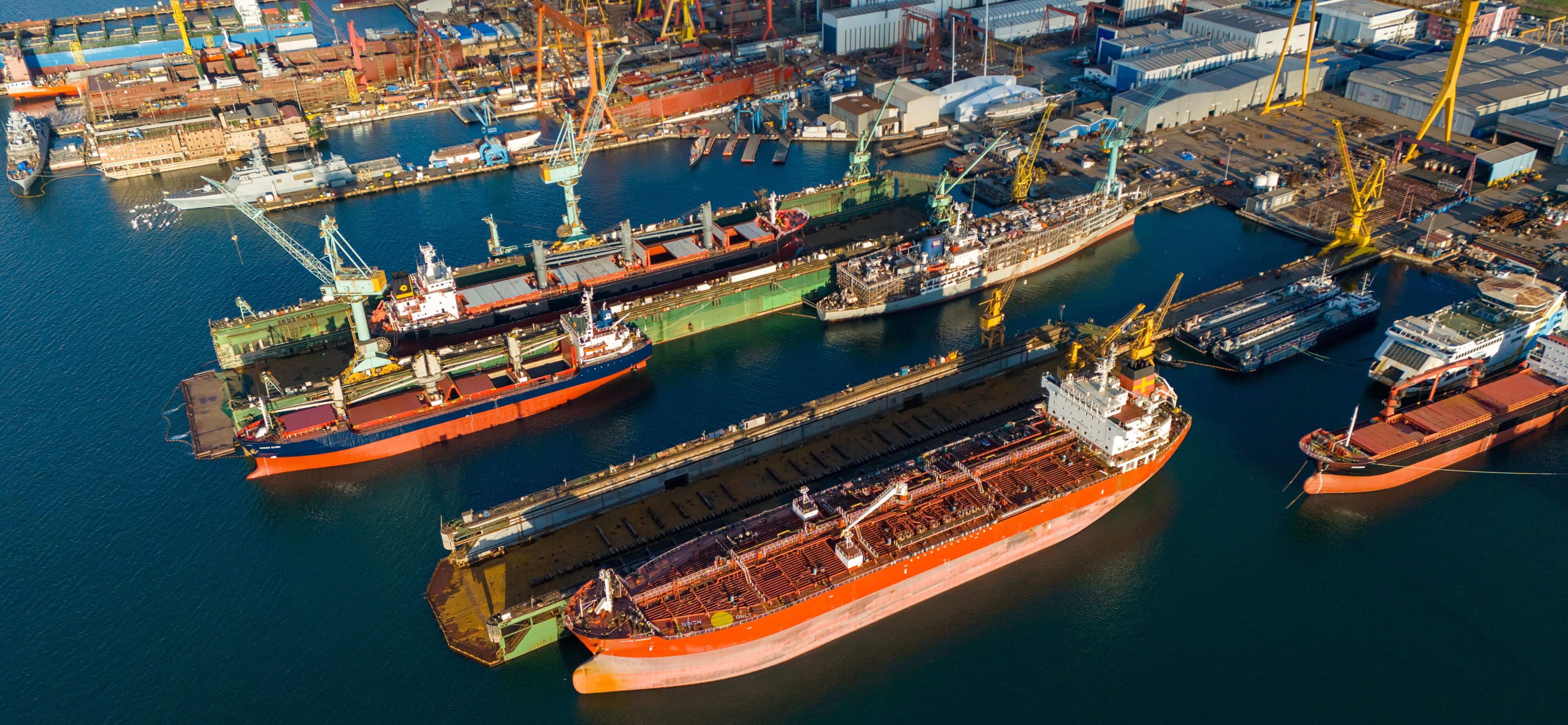 Side view drone shot of a large orange tanker ship lifted in a floating dry dock for hull inspection and painting surrounded by other docked industrial vessels