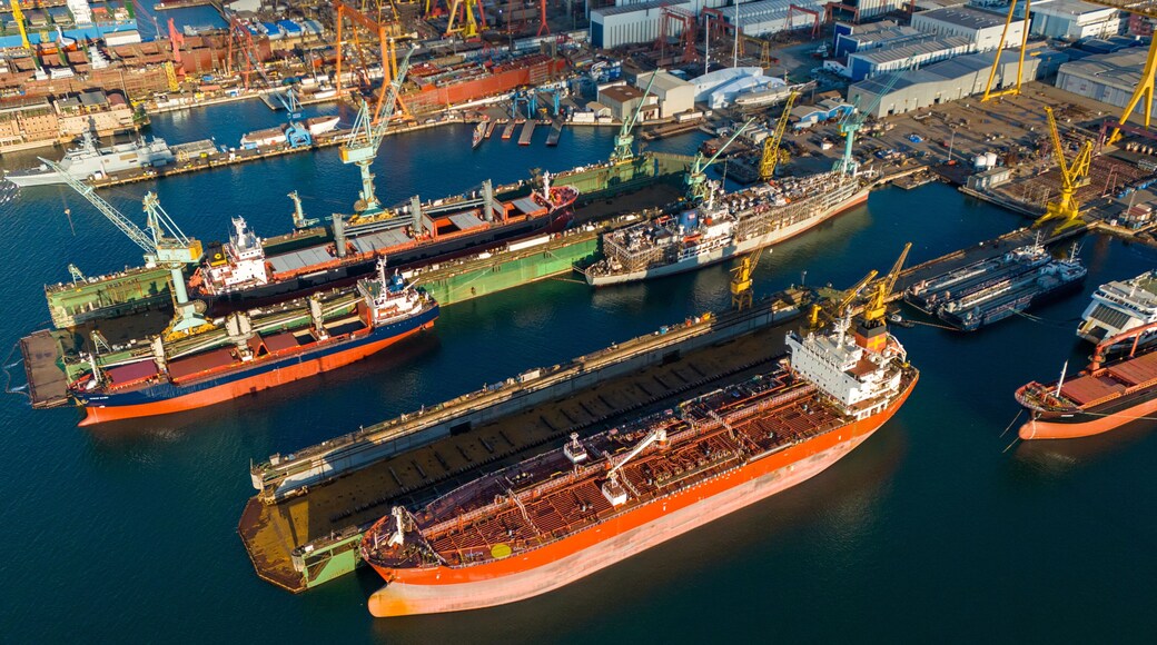Side view drone shot of a large orange tanker ship lifted in a floating dry dock for hull inspection and painting surrounded by other docked industrial vessels