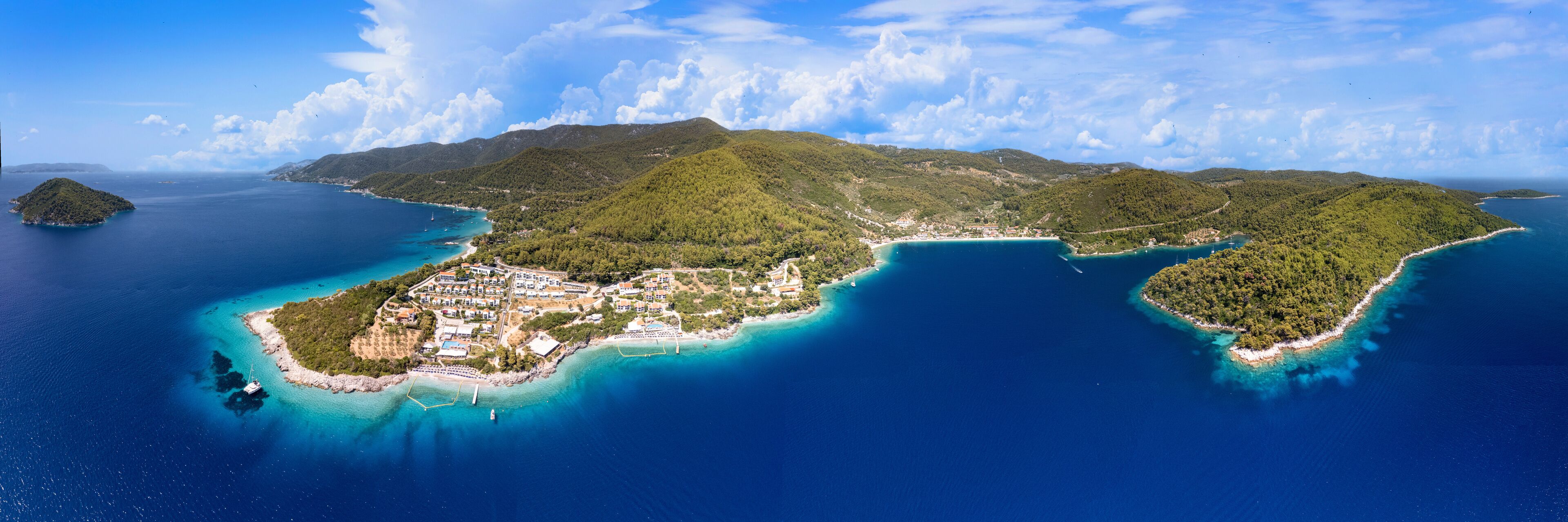 An aerial view of a coastal island with a dense forest, clear blue waters, and a small settlement with buildings and boats.