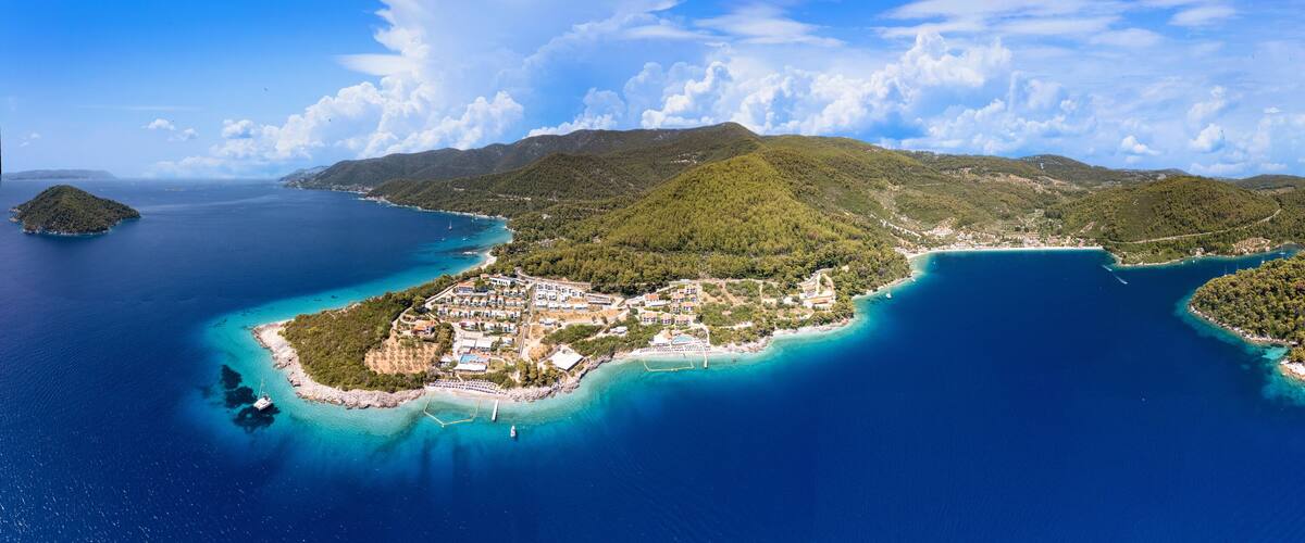 An aerial view of a coastal island with a dense forest, clear blue waters, and a small settlement with buildings and boats.