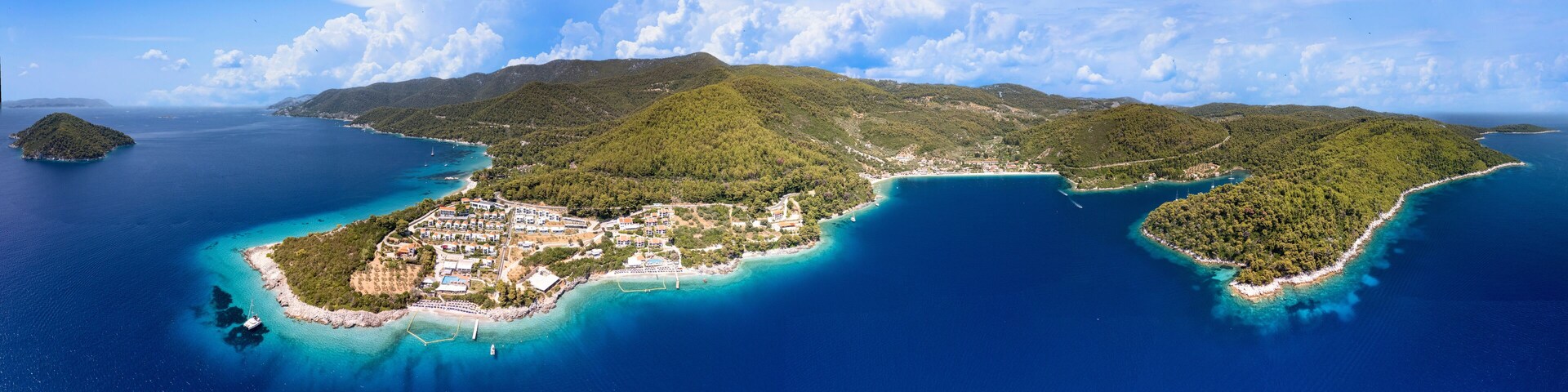 An aerial view of a coastal island with a dense forest, clear blue waters, and a small settlement with buildings and boats.