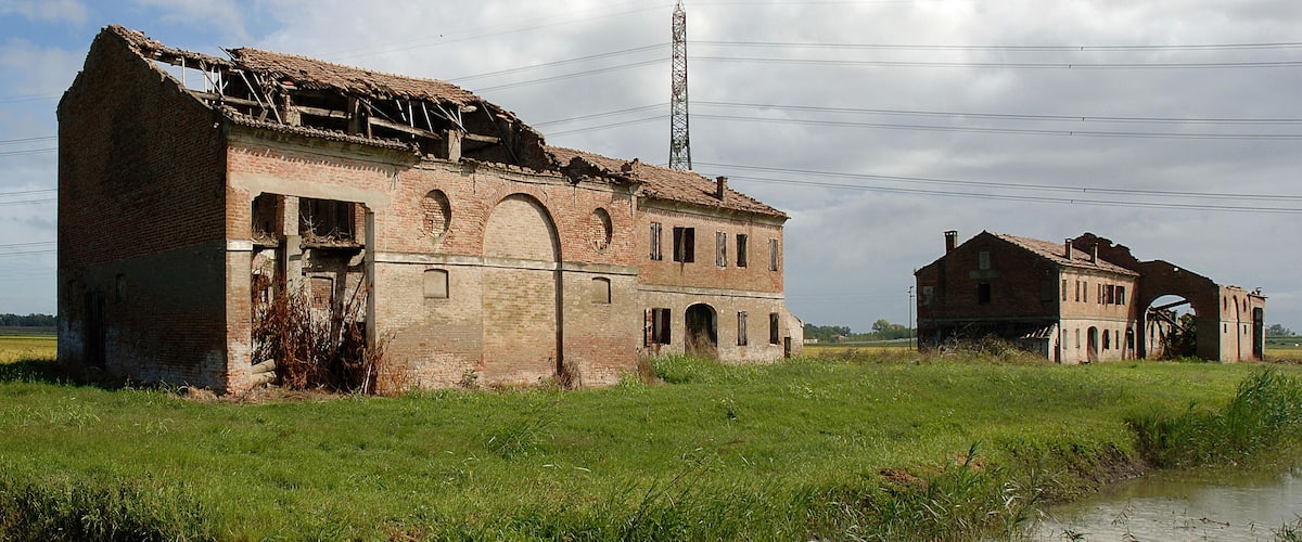 A dilapidated brick building with a collapsed roof, surrounded by a grassy field and a body of water.
