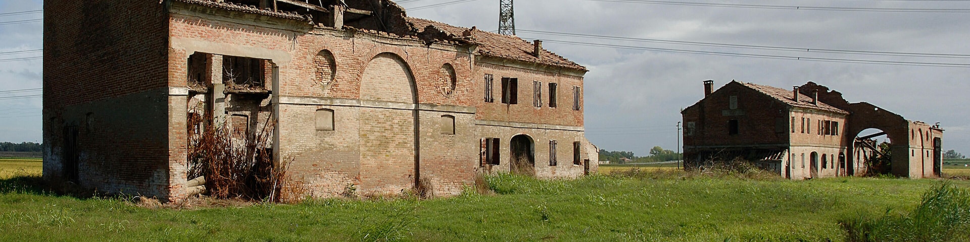 A dilapidated brick building with a collapsed roof, surrounded by a grassy field and a body of water.