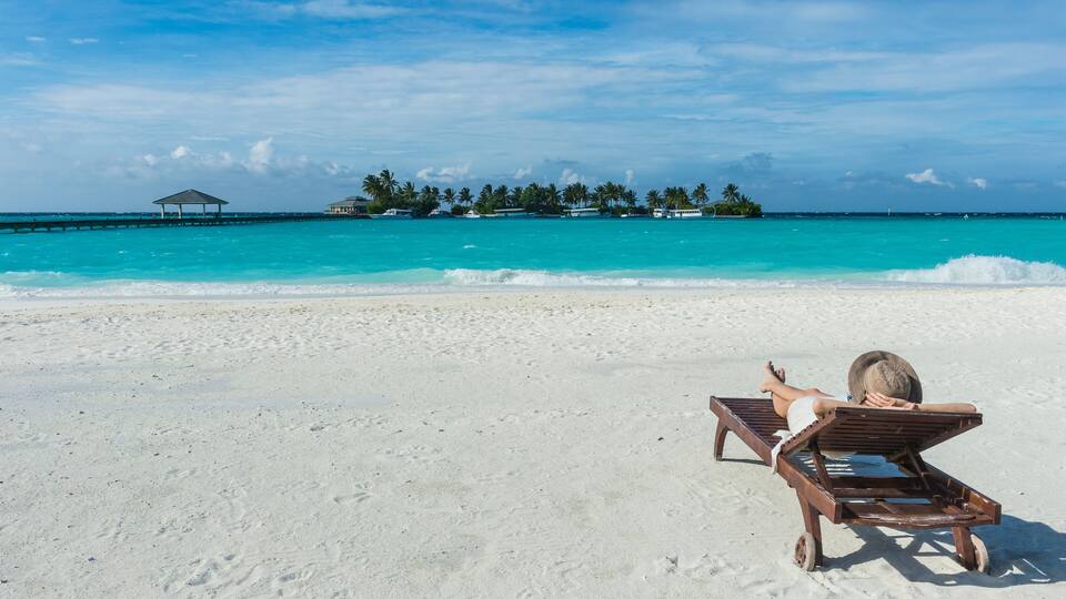A beach with a person lying on a lounge chair, turquoise water, and a small island with palm trees in the background.