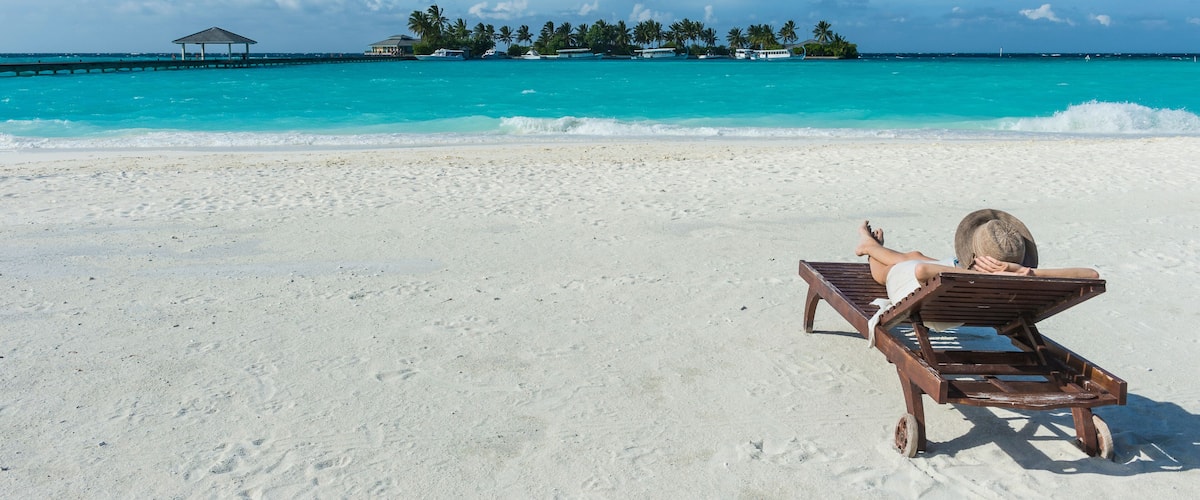 A beach with a person lying on a lounge chair, turquoise water, and a small island with palm trees in the background.
