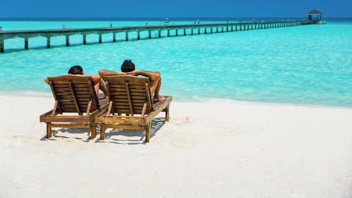 Couple enjoying holidays sitting on lounge chairs at tropical paradise beach at Dhiffushi Holiday island at South Ari atoll, Maldives. Beautiful turquoise Indian ocean sea with white sand. Luxury trav