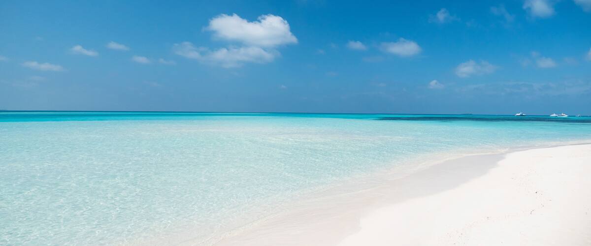 A beach with clear turquoise water and a white sandy shore under a blue sky with scattered clouds.
