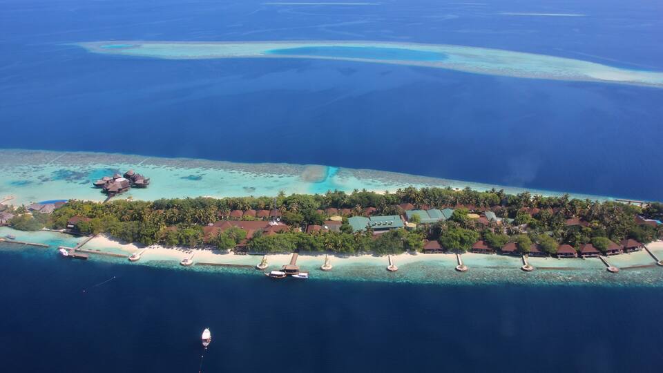 An aerial view of a tropical island with clear turquoise waters, a sandy beach, and overwater bungalows.