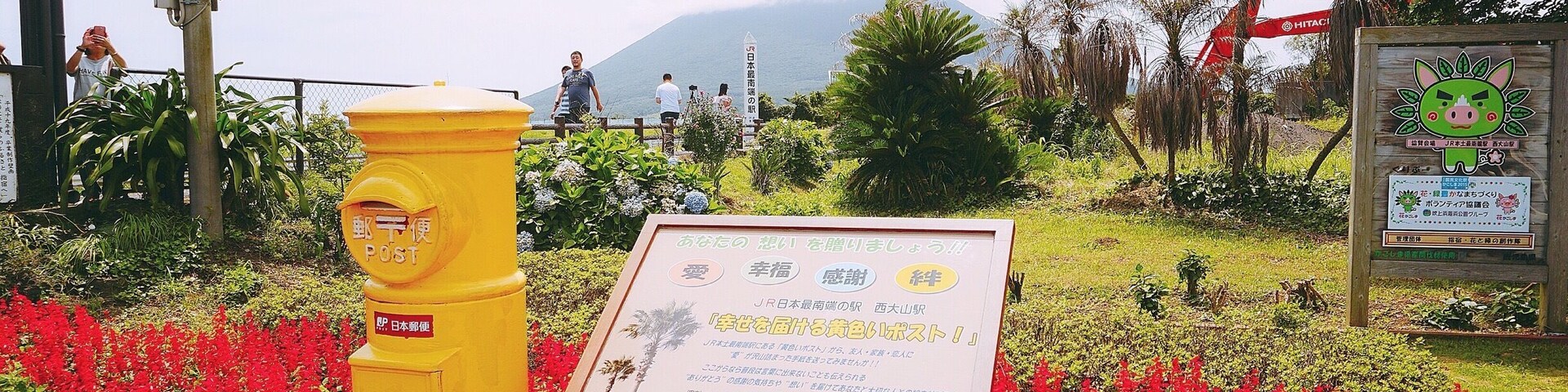 Un hidrante amarillo, un cartel con texto japonés y flores rojas en un jardín con una montaña al fondo.