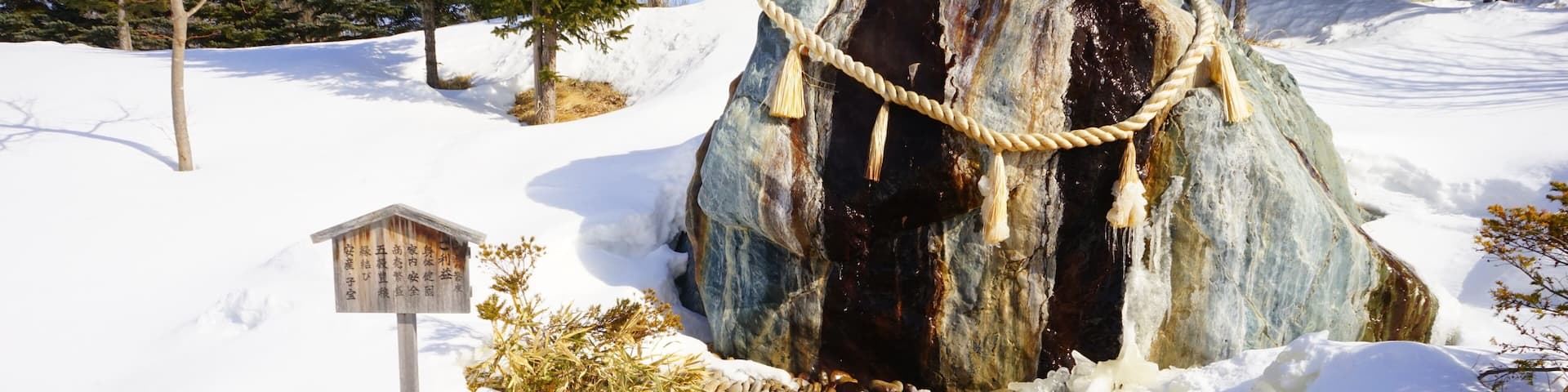 A snowy landscape with a wooden sign, a rope around a rock, and a stream.