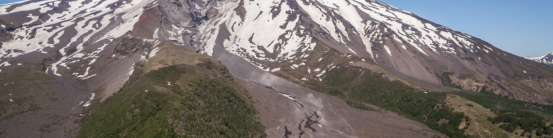 Una montaña nevada con un pico destacado, rodeada de vegetación verde y terreno rocoso.