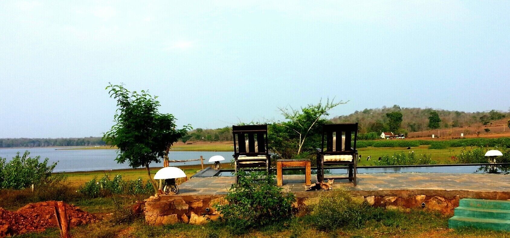 A lakeside seating area with wooden chairs and a small table, surrounded by greenery and a distant view of a hill.