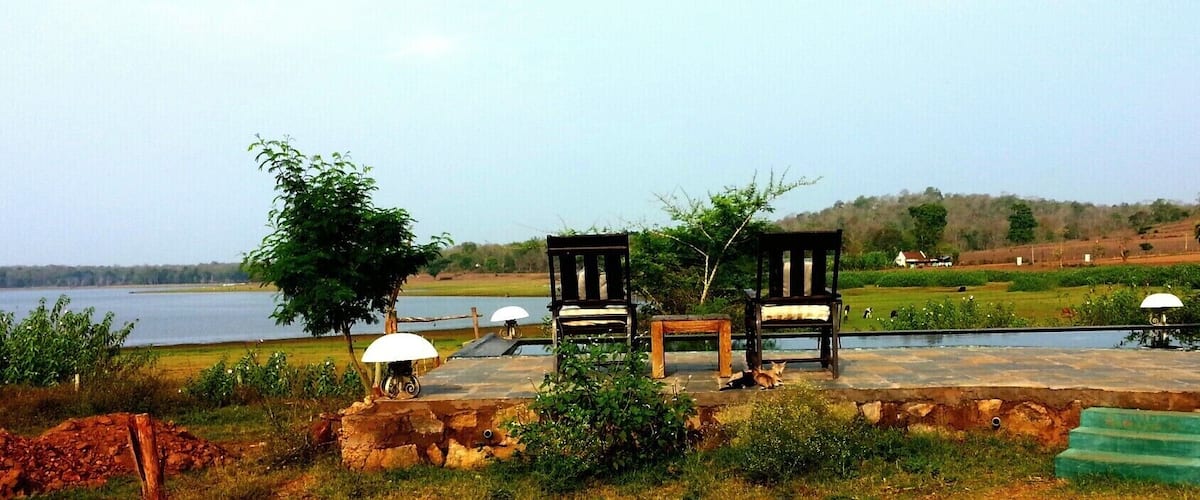 A lakeside seating area with wooden chairs and a small table, surrounded by greenery and a distant view of a hill.