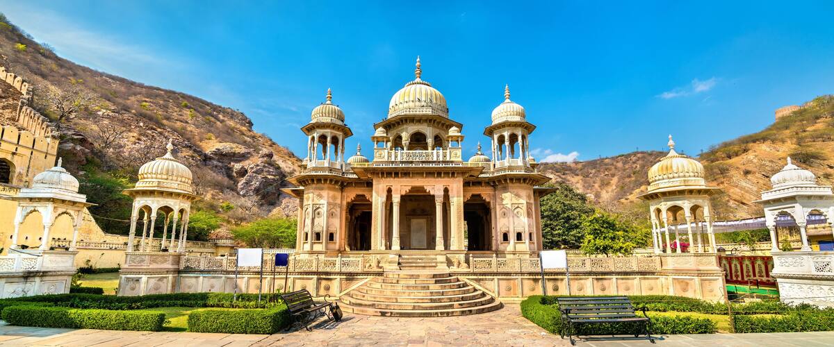 A historic building with multiple domes and intricate carvings, surrounded by a well-maintained garden and a mountainous backdrop.