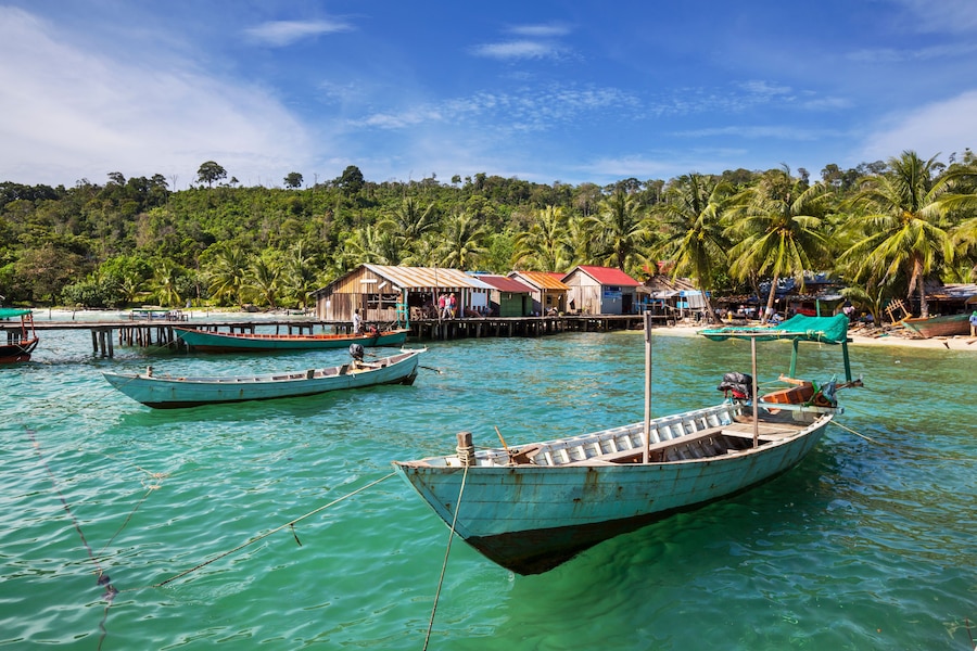 A tropical beach with boats, a wooden pier, and a forested hill.