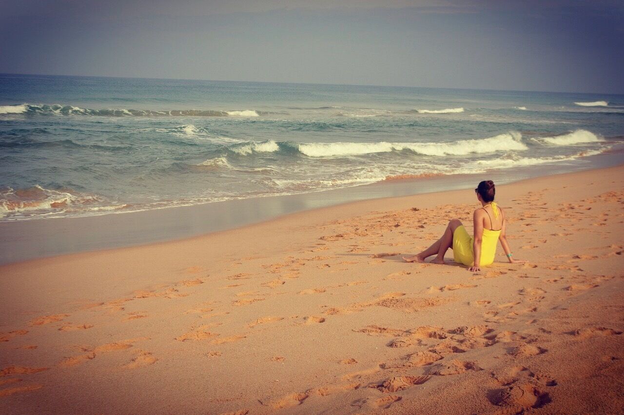 A woman in a yellow dress sitting on a sandy beach with waves in the background.