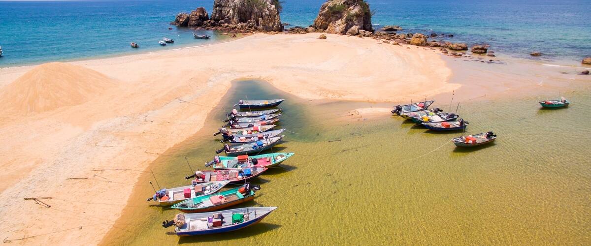 A beach with boats lined up near the shore, clear turquoise water, and rocky formations in the background.