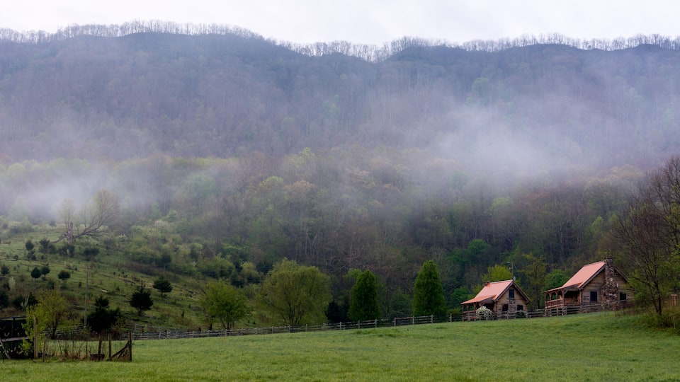 A misty valley with rolling hills, a wooden fence, and two rustic barns.