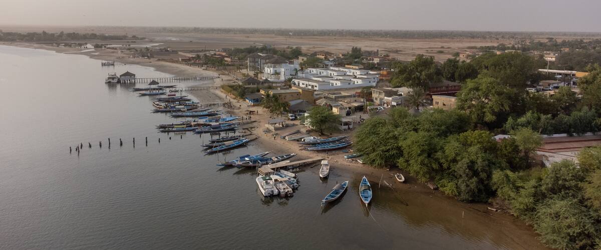 A riverside town with boats docked along the shore, surrounded by buildings and greenery.