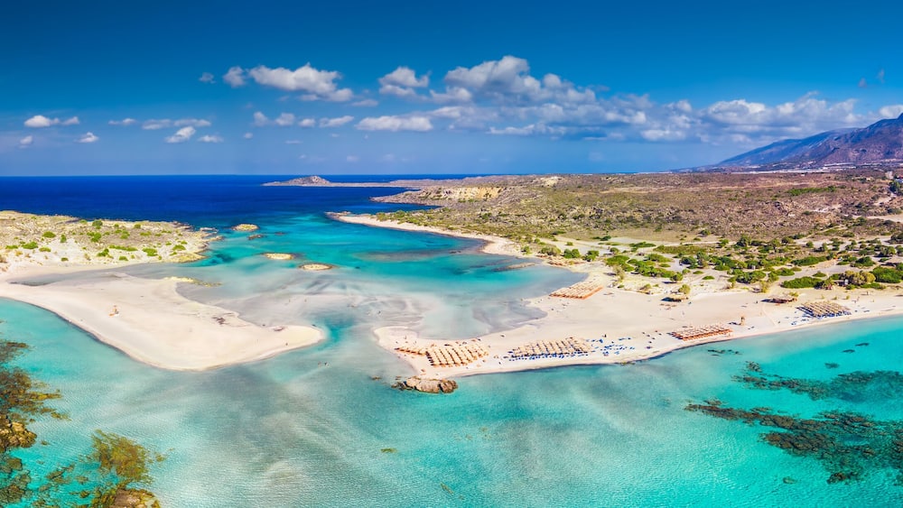 Vue aérienne d’une zone côtière avec des eaux turquoise limpides, des plages de sable et un paysage montagneux en arrière-plan.