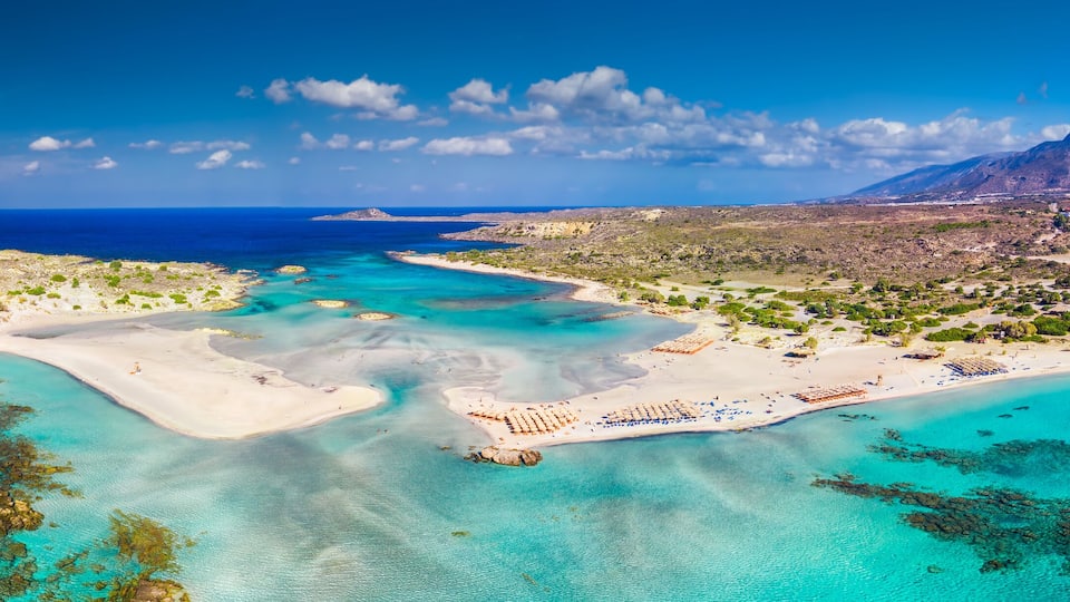 An aerial view of a coastal area with clear turquoise waters, sandy beaches, and a mountainous backdrop.