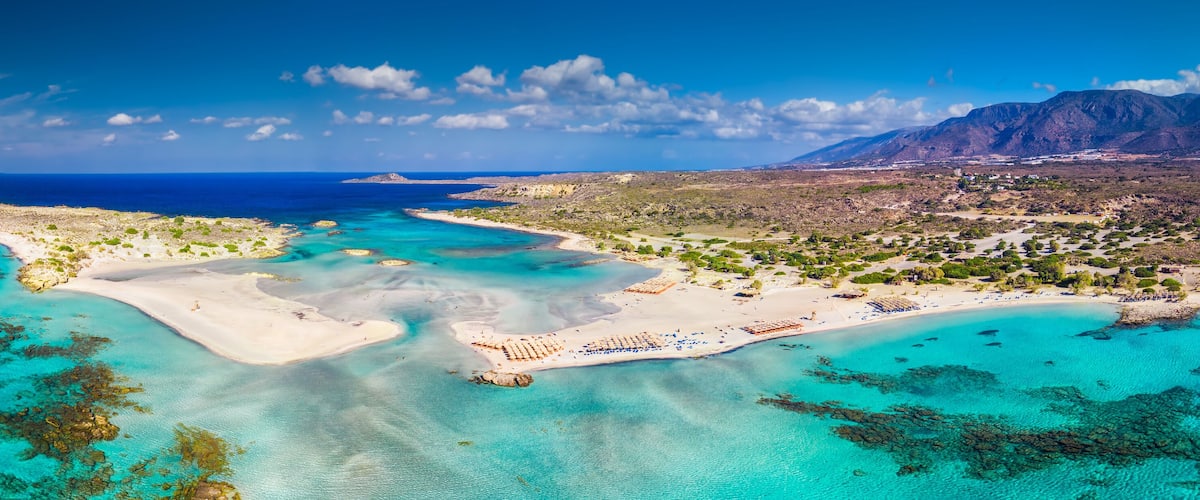An aerial view of a coastal area with clear turquoise waters, sandy beaches, and a mountainous backdrop.