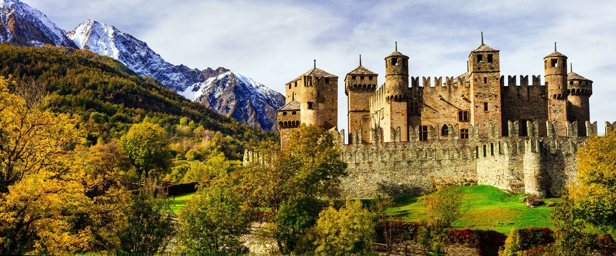 A medieval castle surrounded by autumn foliage and snow-capped mountains.