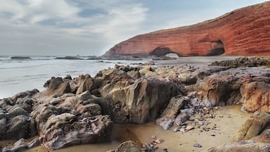 A rocky beach with large boulders and a red cliff with an arch formation.