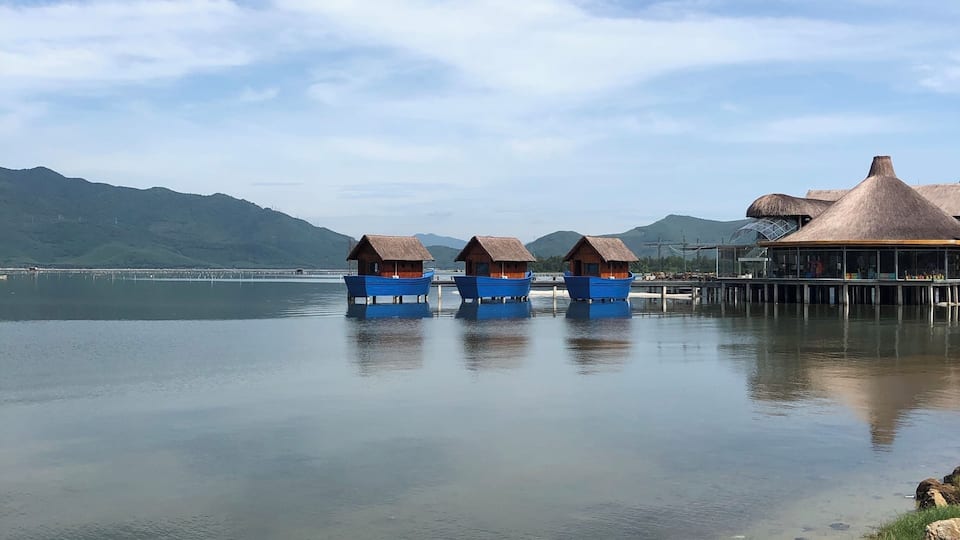 Overwater bungalows on stilts, a calm lake, and mountains in the background.