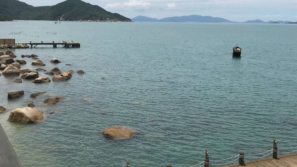 A wooden pier extending into calm waters with scattered rocks, a small boat, and distant mountains under a cloudy sky.