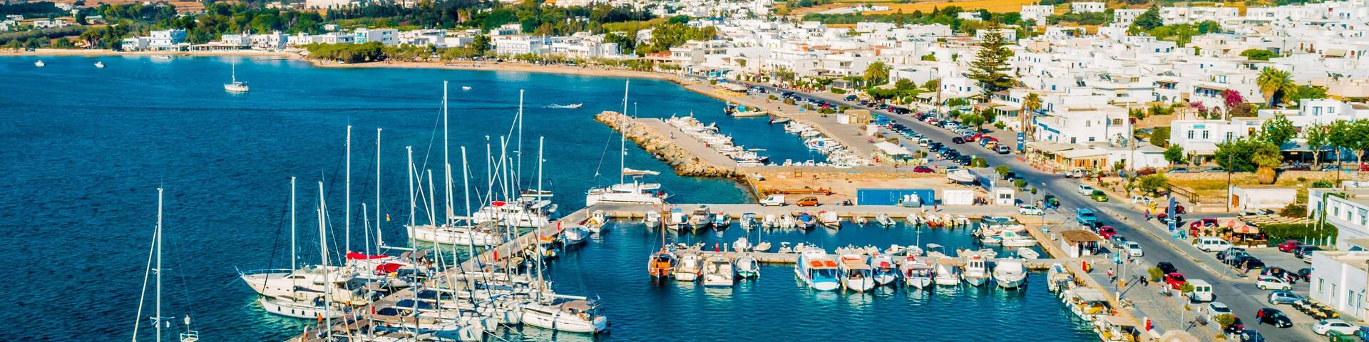 A coastal town with a marina filled with boats, a road along the coast, and hills in the background.