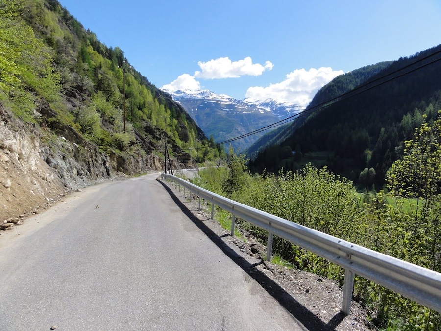 Une route de montagne bordée d’une barrière de sécurité, entourée d’une végétation luxuriante et, au loin, de sommets enneigés.