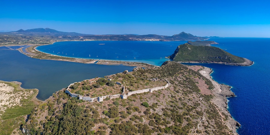 Vista aérea de una región costera con una fortaleza destacada, una amplia bahía con barcos y montañas al fondo.