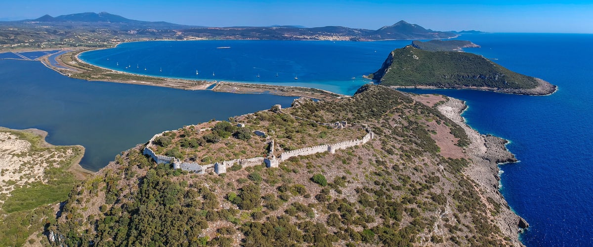An aerial view of a coastal region with a prominent fortification, a large bay with boats, and distant mountains.