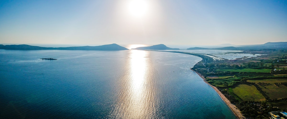 A coastal landscape with a large body of water reflecting sunlight, a distant shoreline, and a hilly background.