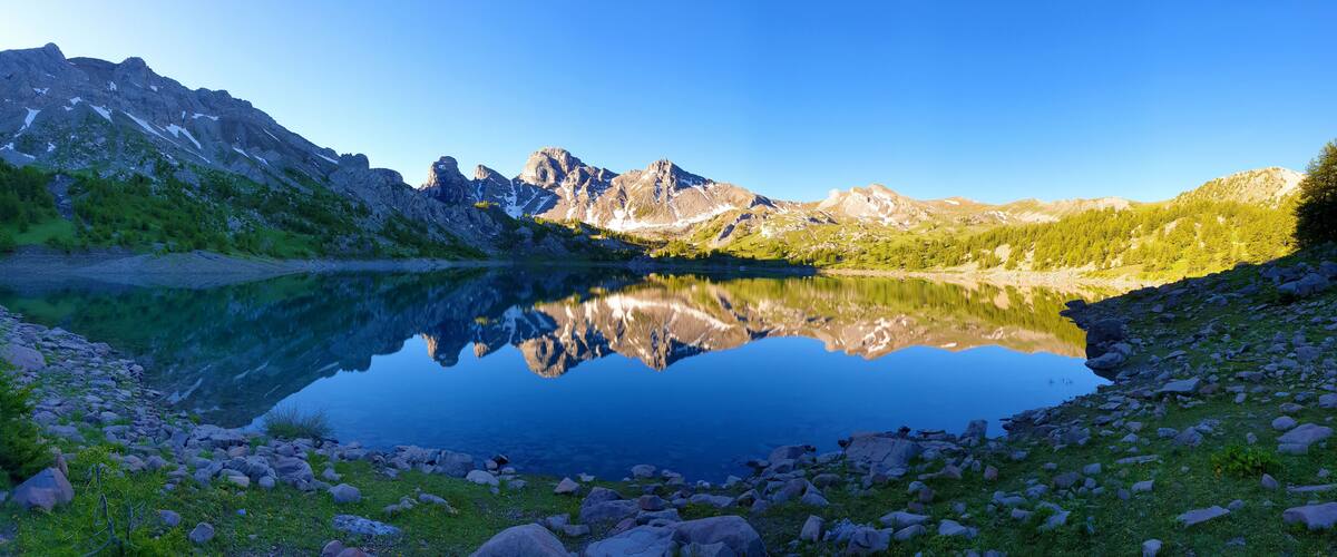 A mountain lake with clear reflections, surrounded by rocky terrain and sparse vegetation.