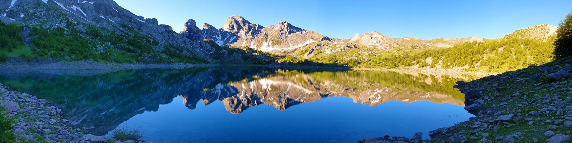 A mountain lake with clear reflections, surrounded by rocky terrain and sparse vegetation.