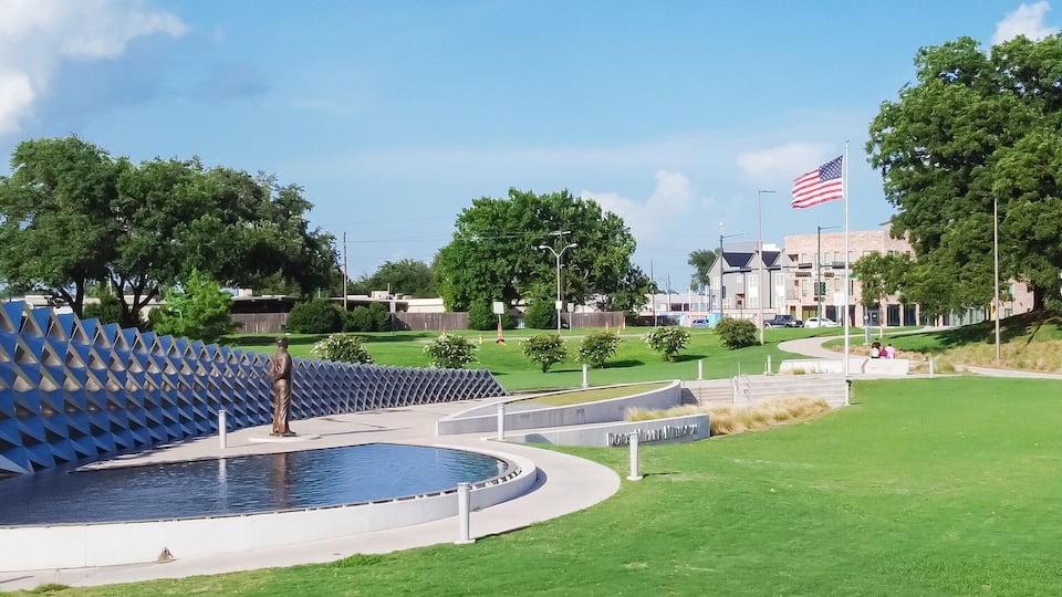 A sculpture of a figure with a blue, geometric structure behind it, a flag, and a grassy park area.
